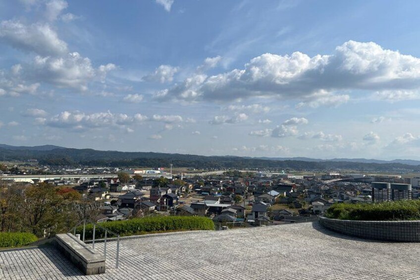 A cityscape with a mix of residential and commercial buildings, a paved area with a curved bench, and distant hills under a partly cloudy sky. 5