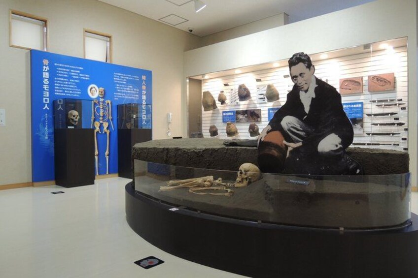 A museum exhibit with a skeleton, a seated figure, and various artifacts on display. 4