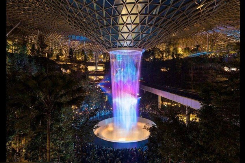 A large indoor waterfall with a circular pool, surrounded by dense greenery and a high, geometric ceiling structure. 1