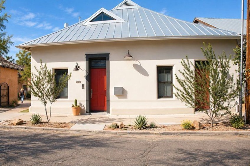 A single-story house with a red door, a metal roof, and small desert plants in front. 3
