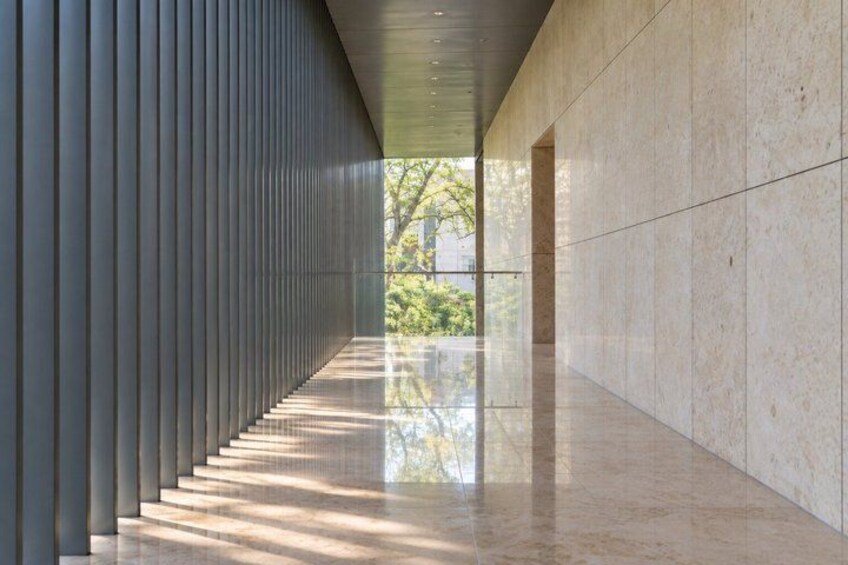 A corridor with a reflective floor, vertical slats on one wall, and a view of greenery outside. 3