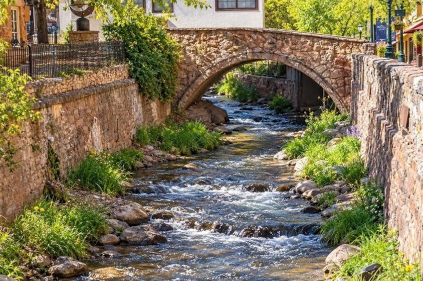 A stone bridge over a flowing river, surrounded by greenery and buildings. 1