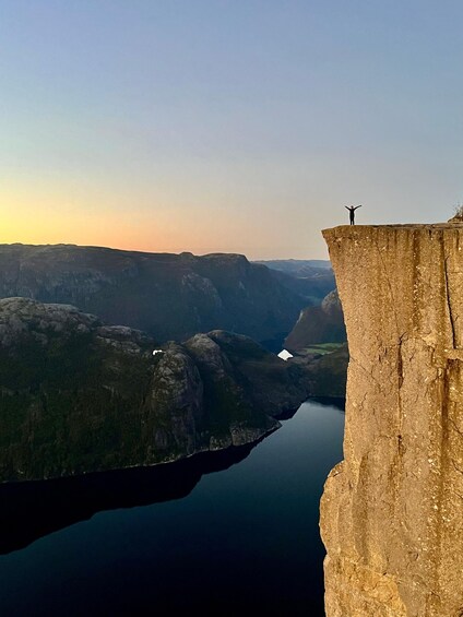 A person standing on a cliff edge with arms outstretched, overlooking a valley and a body of water below. 1