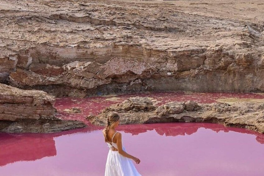 Une personne vêtue d’une robe blanche, debout près d’une piscine rouge, avec un paysage rocheux en arrière-plan. 1