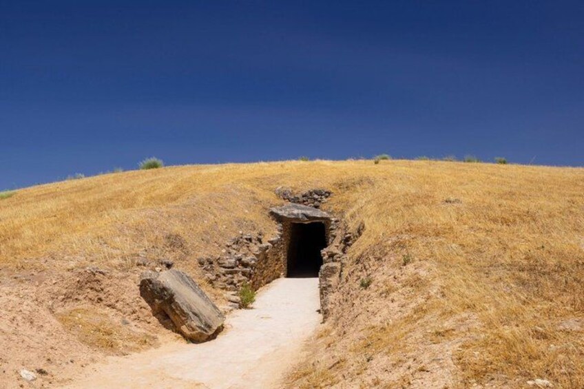 A narrow passageway leading into a cave, surrounded by dry grass and a clear blue sky. 2