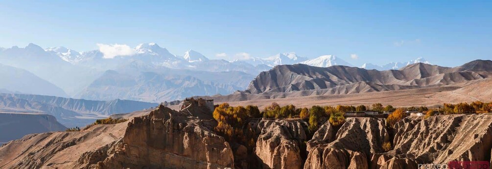 Paisaje montañoso con un monasterio en un acantilado, picos nevados al fondo y árboles de colores otoñales. 3