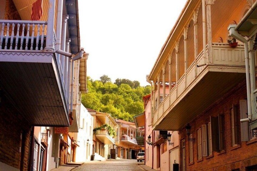 A narrow street flanked by buildings with balconies, a car parked on the right, and a hill with greenery in the background. 1
