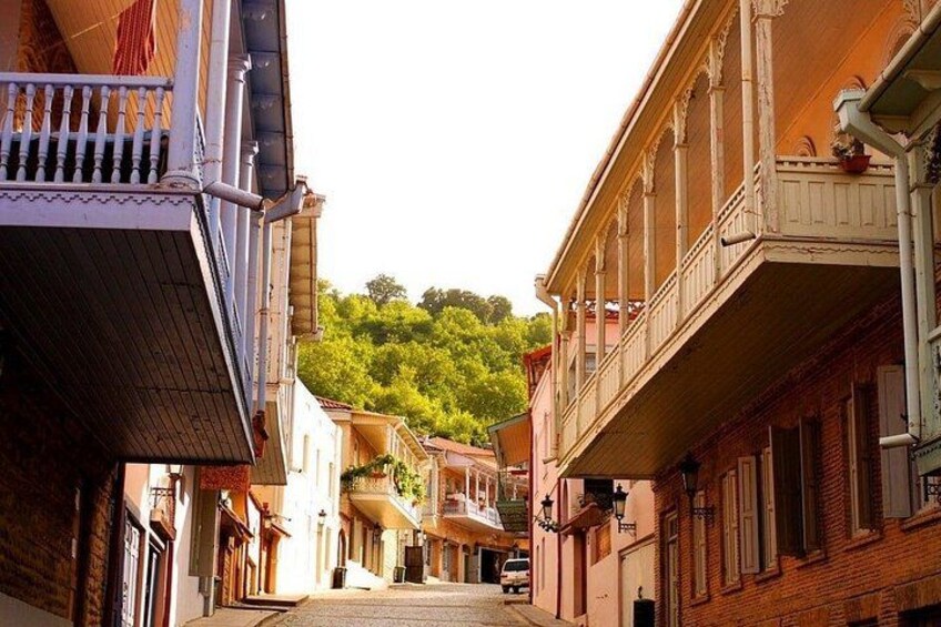 A narrow street flanked by buildings with balconies, a car parked on the right, and a hill with greenery in the background. 1