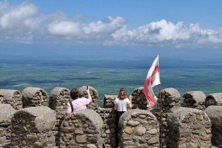 A stone wall with a flag, a person taking a photo, and a view of the ocean. 3