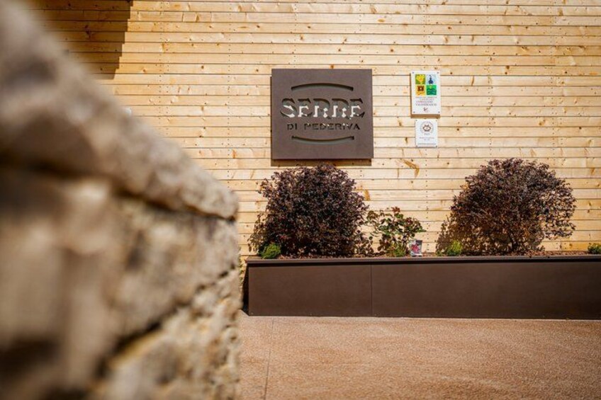 A wooden wall with a sign that reads 'SERRA DI MENERA' and two small bushes. 2