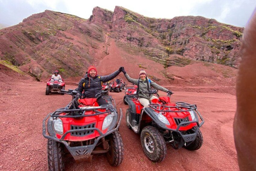 Two people on red ATVs in a red rocky terrain with hills in the background. 3