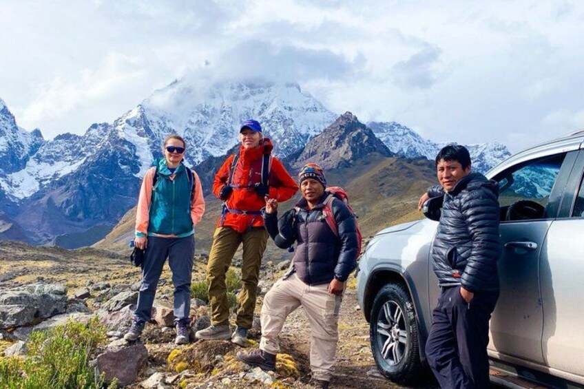 Four people standing on a rocky terrain with a mountainous background and a parked vehicle. 1