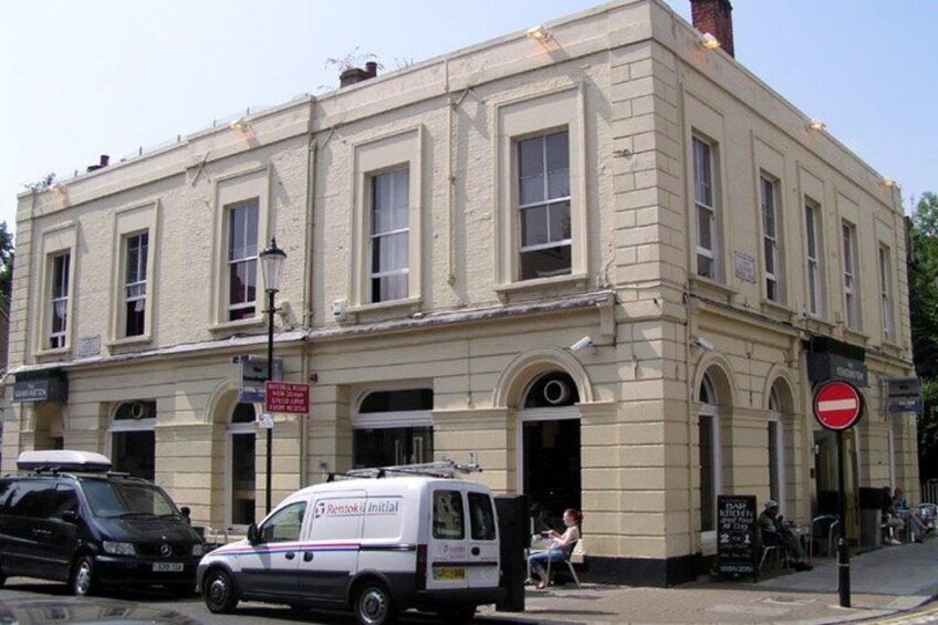A two-story building with arched windows and a signboard. A white van is parked in front, and a black car is visible on the left. 5