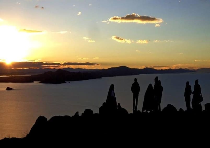 Ein Sonnenuntergang über einem Gewässer mit Silhouetten von Menschen, die an einem felsigen Ufer stehen. 4