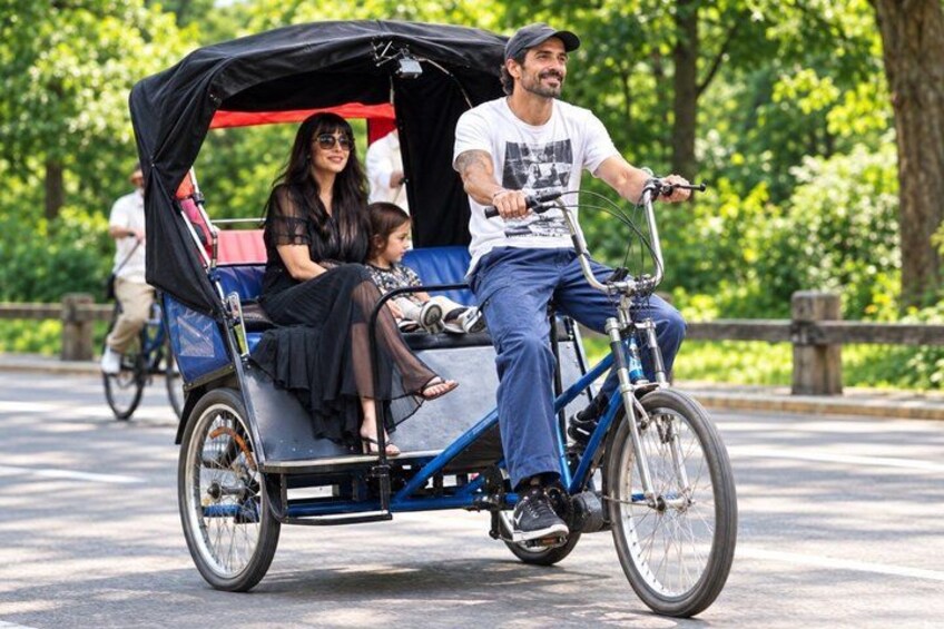 A three-person rickshaw with a black canopy, a man driving, and two passengers. 1