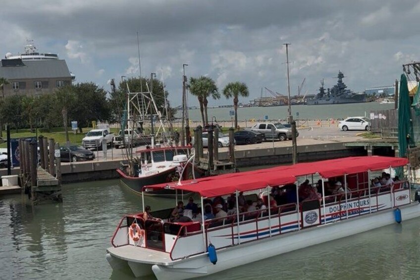 A boat tour with a red canopy, docked at a marina with other boats and a large ship in the background. 5