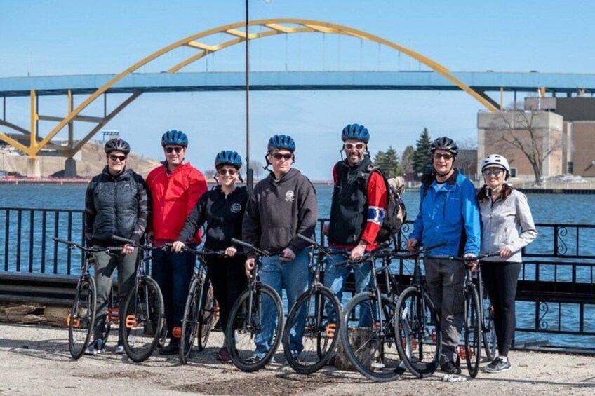 A group of people with bicycles by a river with a bridge in the background. 1