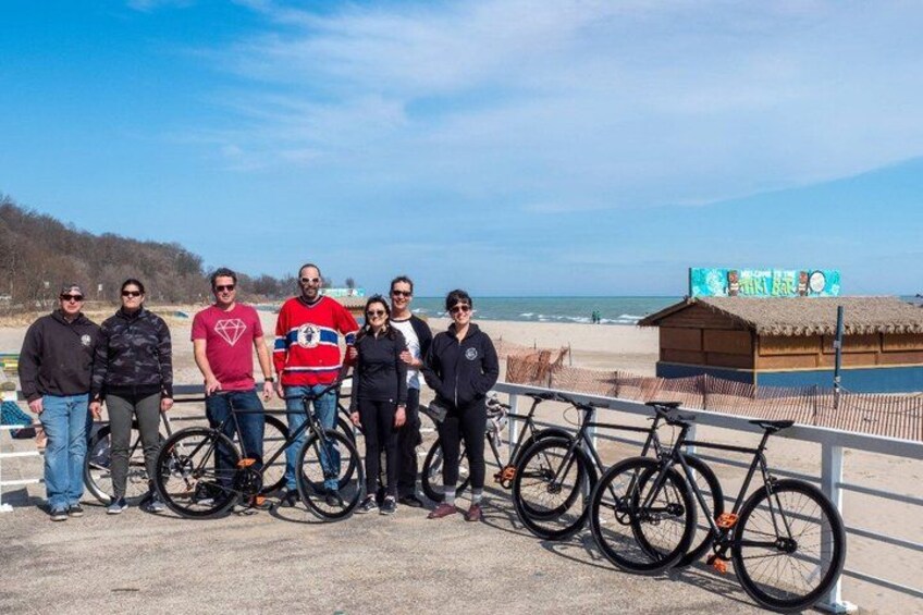 A group of people standing by bicycles on a beach with a thatched-roof structure in the background. 2