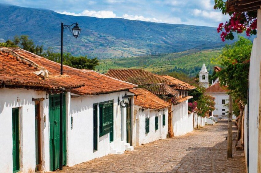 A cobblestone street lined with white buildings with green shutters, a church, and mountains in the background. 1