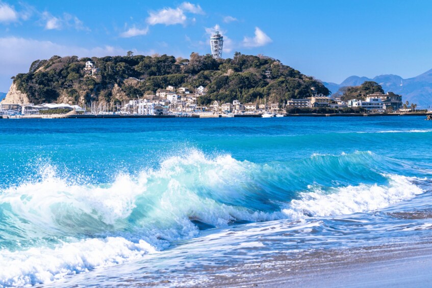 A coastal town with a prominent lighthouse on a hill, clear blue waters, and waves crashing onto the shore. 2