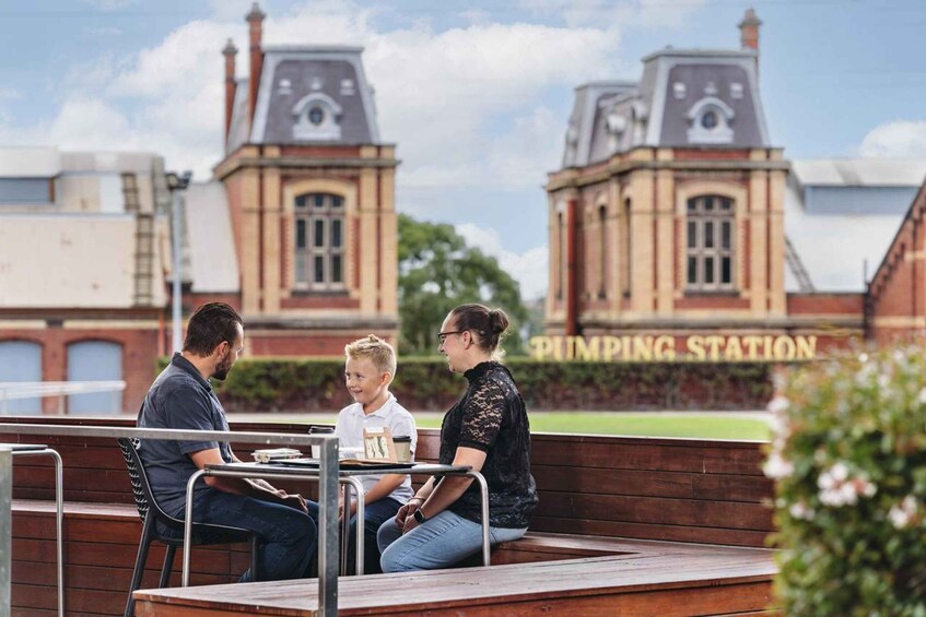 Un groupe de personnes est assis à une table à l’extérieur, et on aperçoit en arrière-plan un bâtiment historique portant l’inscription « STATION DE POMPAGE ». 1