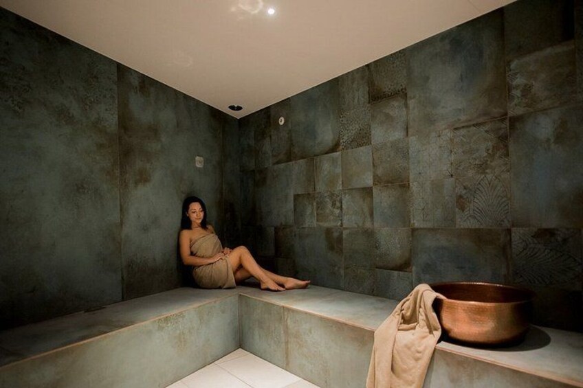 A person sitting on a bench in a room with textured walls and a copper pot on a ledge. 2