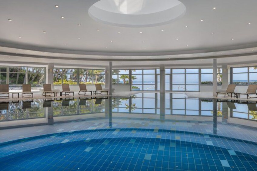 An indoor swimming pool with clear blue water, surrounded by lounge chairs and large windows overlooking the ocean. 1