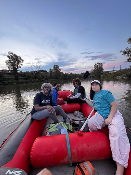 A group of people in a red inflatable raft on a calm river with trees and a bridge in the background. 3