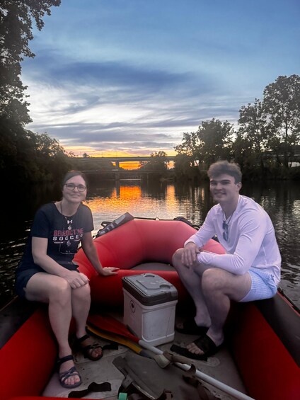 Two people sitting in a red inflatable boat on a calm river with a sunset in the background. 4