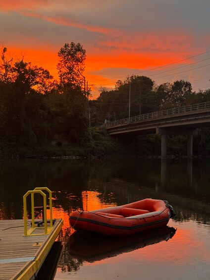 A sunset over a river with a red inflatable boat docked at a wooden pier. 1