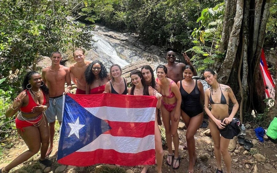 Un grupo de personas sostiene una bandera de Puerto Rico en un área boscosa con una cascada al fondo. 1