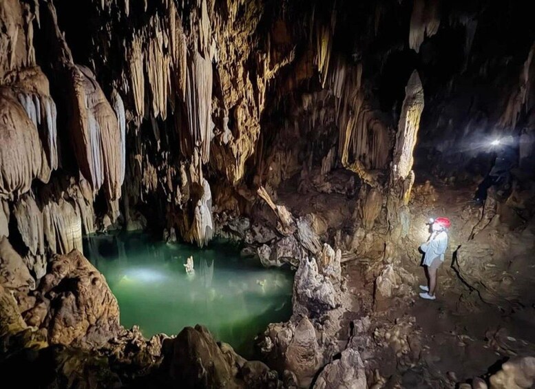 A cave with stalactites and stalagmites, a person wearing a helmet, and a green pool of water. 1