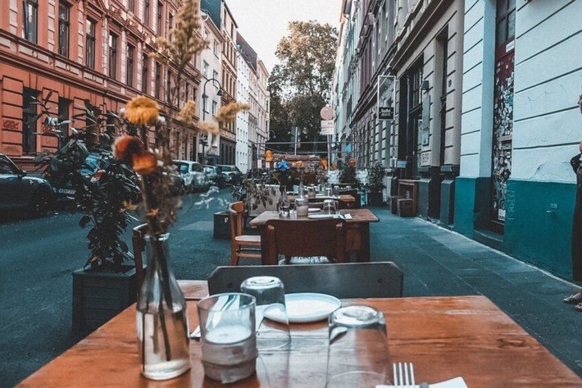 An outdoor dining area with tables set for two, surrounded by buildings and parked cars. 3