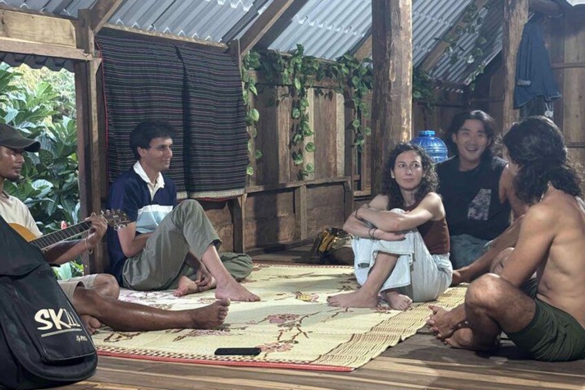 A group of people sitting on a mat in a rustic outdoor setting with wooden structures and greenery in the background. 2