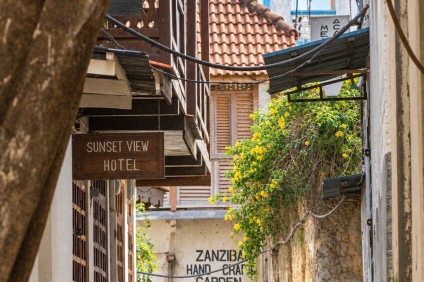 A narrow street with buildings, a sign for 'Sunset View Hotel,' and a wall with 'Zanzibar Handcraft Garden' written on it. 3