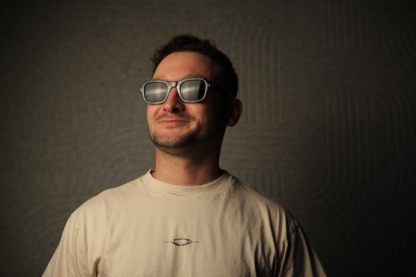 A person wearing a white t-shirt with a small logo on the chest, standing against a textured dark background. 4