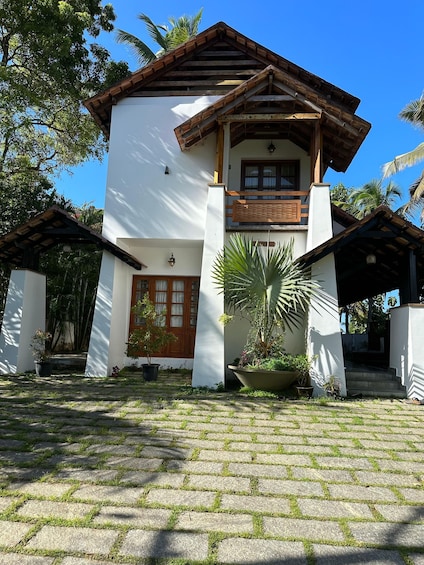 A two-story house with a white facade and wooden accents, surrounded by lush greenery and palm trees. 1