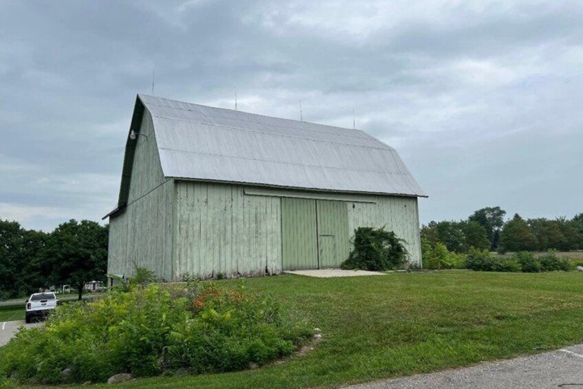 A large, single-story barn with a sloped roof, surrounded by a grassy field and a few trees in the background. 4