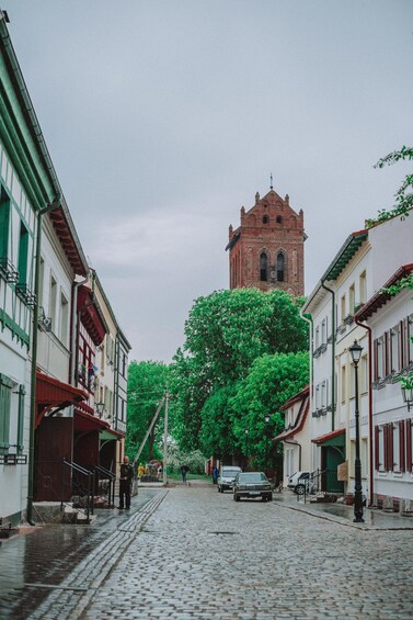 A cobblestone street lined with historic buildings, a tall brick tower, and lush green trees. 2