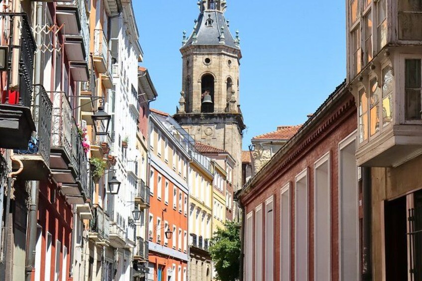 A narrow street lined with colorful buildings, a prominent clock tower, and a clear blue sky. 1