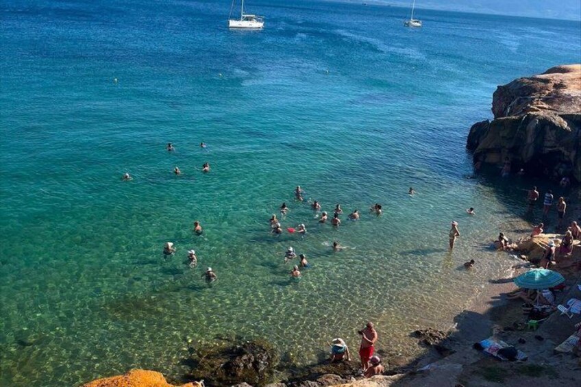 A beach with people swimming and sunbathing, clear turquoise water, and rocky outcrops. 4