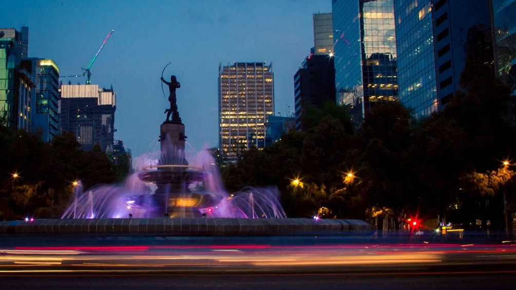 Un paisaje urbano al anochecer con una fuente en primer plano y edificios iluminados al fondo. 1