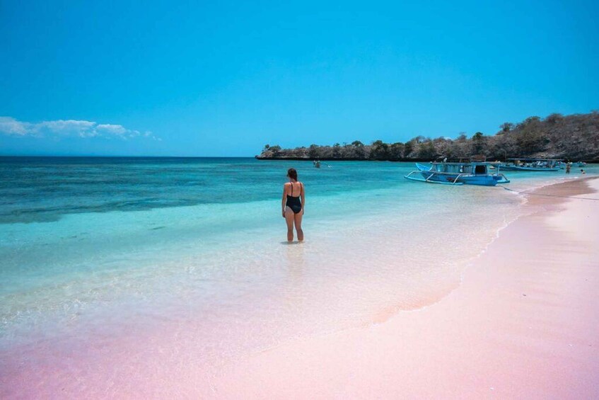 A woman in a swimsuit standing on a beach with clear turquoise water, boats, and a distant island. 3