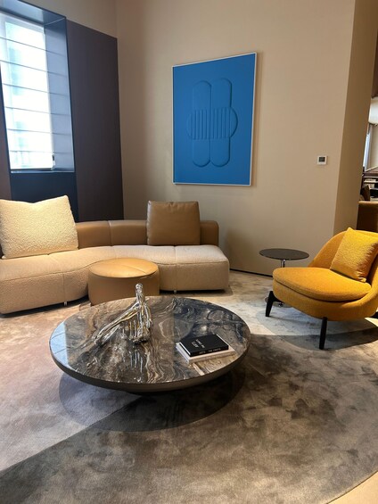 Ein modernes Wohnzimmer mit einem beigefarbenen Ecksofa, einem gelben Sessel, einem runden Couchtisch mit Glasplatte und einem blauen abstrakten Gemälde an der Wand. 3