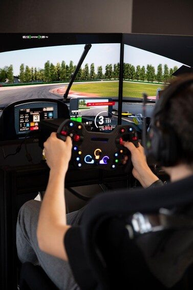 A person driving a racing simulator with a steering wheel and pedals, a dashboard with various gauges and a screen displaying a race track. 3
