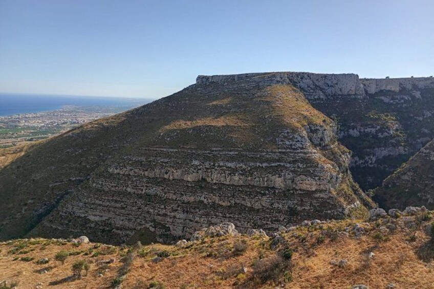 Una gran formación rocosa estratificada con cima plana, rodeada por un paisaje montañoso y una vista distante de un pueblo. 1