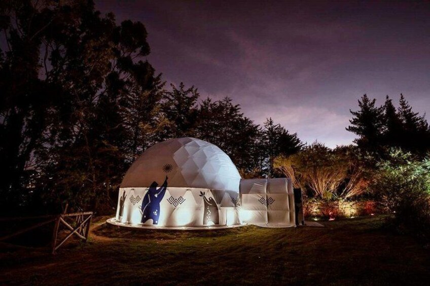A geodesic dome structure at night with a white exterior and a blue interior, surrounded by trees and a grassy area. 2