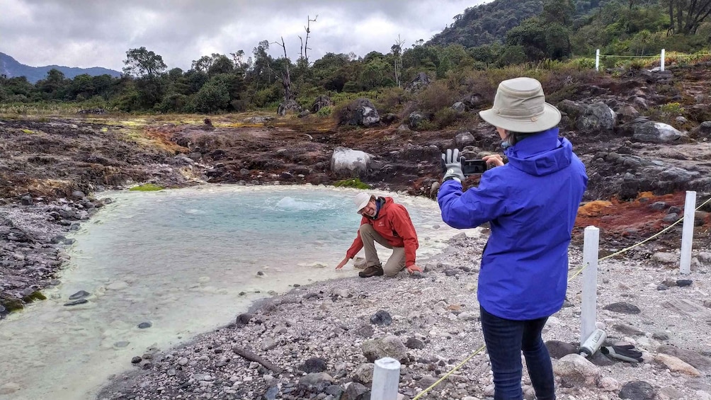 A person in a blue jacket taking a photo of another person in a red jacket near a geothermal pool. 1
