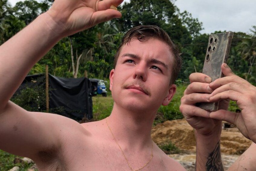 A person holding a smartphone, with a tattoo on their arm, in a natural outdoor setting with trees and a tent in the background. 2
