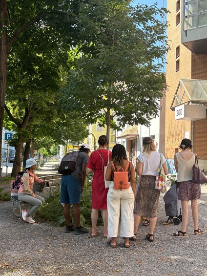 A group of people standing on a gravel path, some with backpacks, near a tree and a building with a number sign. 3